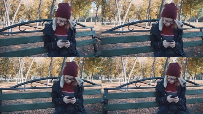 女孩拿着你的手机坐在公园的长椅上。穿着夹克和帽子的女孩