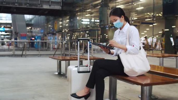 迷人的女商人戴着外科口罩在平板电脑上工作，坐在机场航站楼的椅子上检查电子邮件，并在新型冠状病毒肺炎大