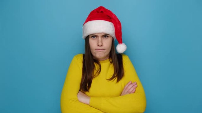 沮丧的不快乐的年轻女子带着沮丧的悲伤表情看着相机，感到被侮辱或冒犯，穿着毛衣和圣诞帽，孤立在蓝色背景