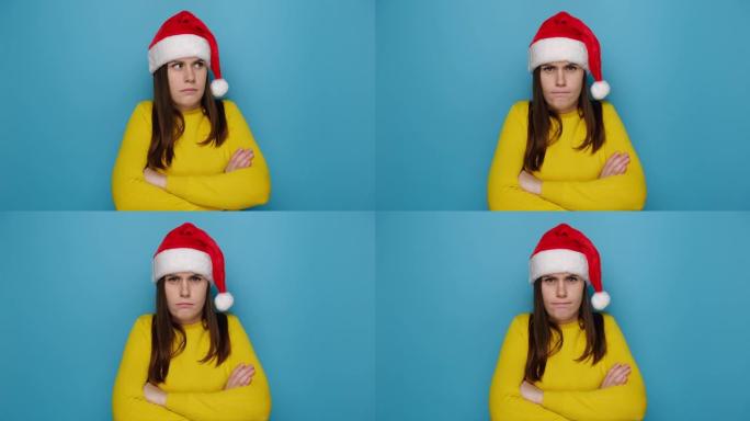 沮丧的不快乐的年轻女子带着沮丧的悲伤表情看着相机，感到被侮辱或冒犯，穿着毛衣和圣诞帽，孤立在蓝色背景