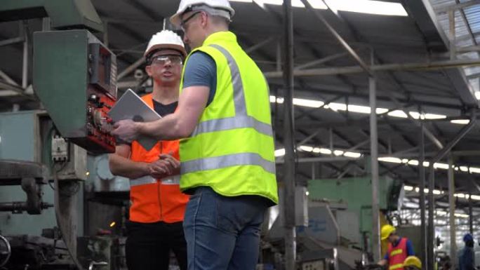 当工人在工业工厂工作时，戴着安全帽的工程师和检查员经理一起交谈，并用平板电脑检查机器。