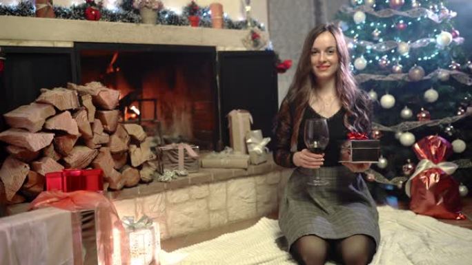 美丽的女人，手里拿着一杯红酒和圣诞节，坐在燃烧的壁炉和圣诞树附近，房间里有令人惊叹的发光装饰，家里有