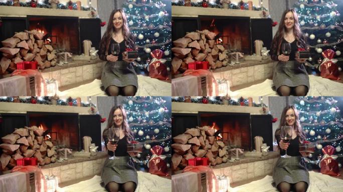 美丽的女人，手里拿着一杯红酒和圣诞节，坐在燃烧的壁炉和圣诞树附近，房间里有令人惊叹的发光装饰，家里有