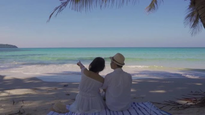 恋爱中的年轻亚洲夫妇坐在热带海滩的棕榈树下，在度蜜月的时候看着美丽的夏日大海。暑假和假期概念。