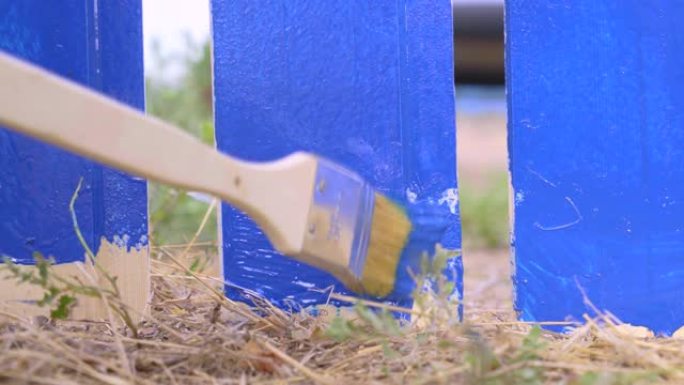 杂工用木栅栏用油漆刷和蓝色染料在村子里画木板底座，特写。地块的维修和建设工作