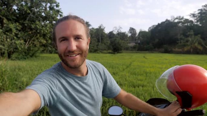 摩托车上的年轻人在日落时在稻田附近拍摄自拍视频，在线分享旅行之旅。人们探索时髦的交通概念