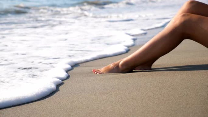 女人的纤细的腿躺在热带海滩上，海浪飞溅。海潮冲刷晒黑的雌性脚。暑假在海岸放松的女孩。暑假概念。多莉开
