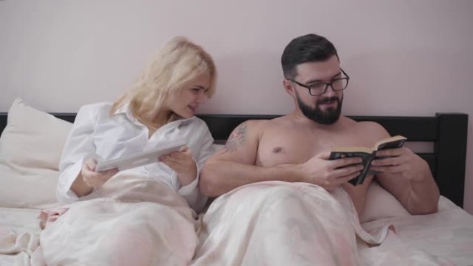 快乐的高加索配偶晚上躺在床上的肖像。可爱的金发女人用平板电脑，男人看书。妻子在看丈夫的故事。家庭讨论