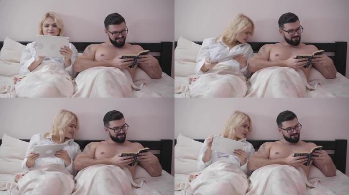 快乐的高加索配偶晚上躺在床上的肖像。可爱的金发女人用平板电脑，男人看书。妻子在看丈夫的故事。家庭讨论
