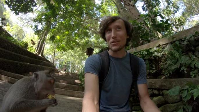 在巴厘岛乌布村的猴子森林自然公园里，一个年轻人与猕猴喂食并自拍的慢动作镜头