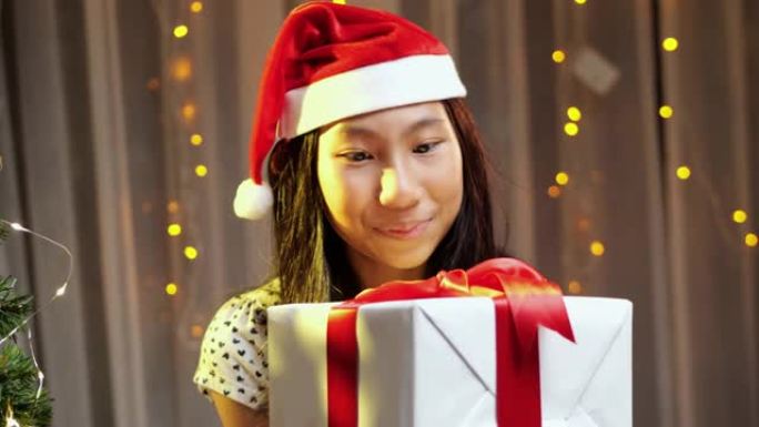 亚洲女孩戴着圣诞老人的帽子，晚上在家拿着带有笑脸的圣诞礼物。