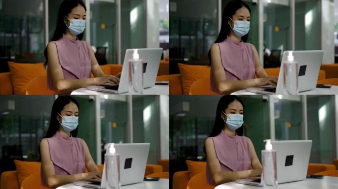 亚洲女商人戴着Covid-19防护口罩在现代办公室工作，使用电脑，专注、防护、健康——新的正常工作生