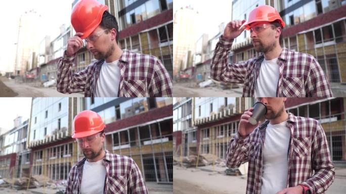 年轻的高加索男性工头建筑商累了，在背景建筑工地喝咖啡。保护设备的工程师咖啡休息时间。辛苦一天后，建筑