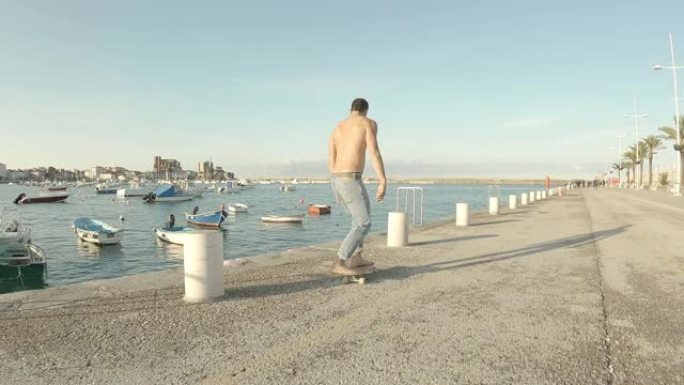 穿着蓝色牛仔裤的年轻人在下午以大海为背景的码头上赤脚光着膀子溜冰
