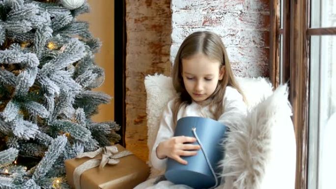 可爱的金发女孩正在打开深蓝色礼品盒，她坐在圣诞树附近，童年快乐和圣诞庆祝概念