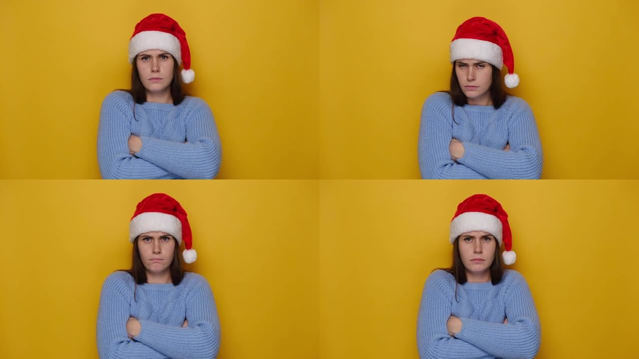 阴郁的受侮辱的年轻女子站在防御姿势，冒犯了鬼脸，等待道歉，戴着圣诞红色帽子和冬季毛衣，在黄色工作室背