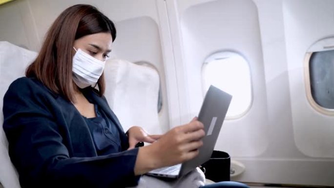 戴着口罩的亚洲妇女使用笔记本电脑，坐在窗户附近的飞机椅子上。健康保障航空旅行运输的概念新常态。