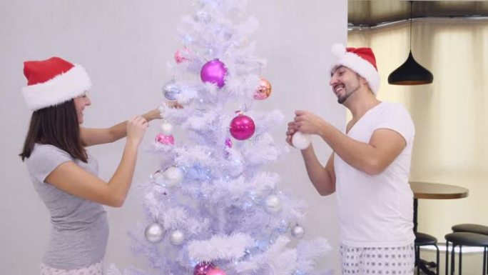 有趣的妻子和丈夫站在银色圣诞树的不同侧面，鬼混和装饰，穿着漂亮的圣诞老人帽子。