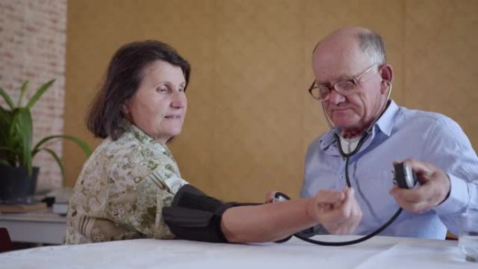有爱心的老人使用血压计测量坐在厨房餐桌上的健康老年妇女的压力，