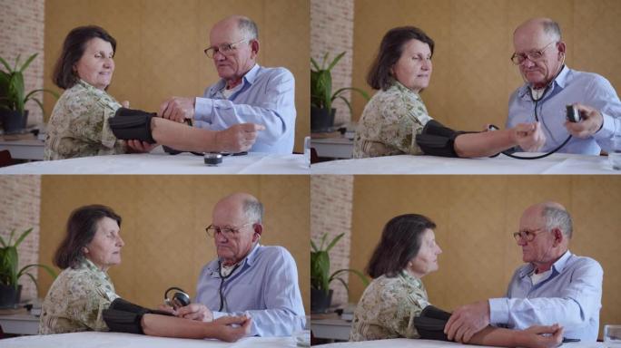 有爱心的老人使用血压计测量坐在厨房餐桌上的健康老年妇女的压力，