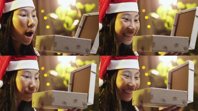 快乐的亚洲女孩戴着圣诞老人的帽子，在家打开带圣诞灯的礼品盒。