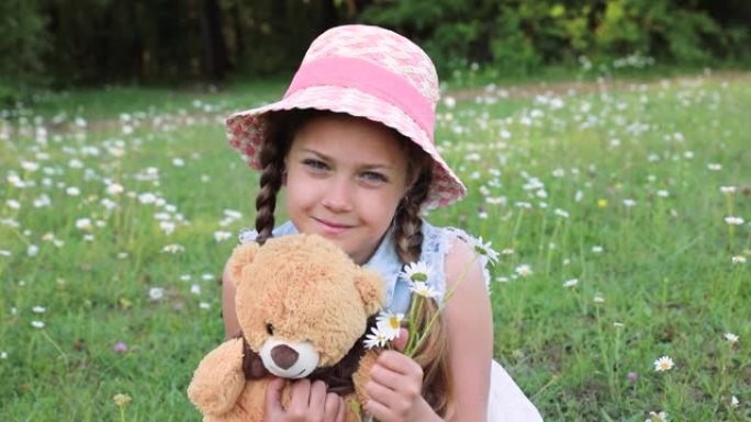 一个戴着帽子的可爱的小女孩在玩她的泰迪熊。婴儿和最喜欢的玩具。夏天，绿色林间空地