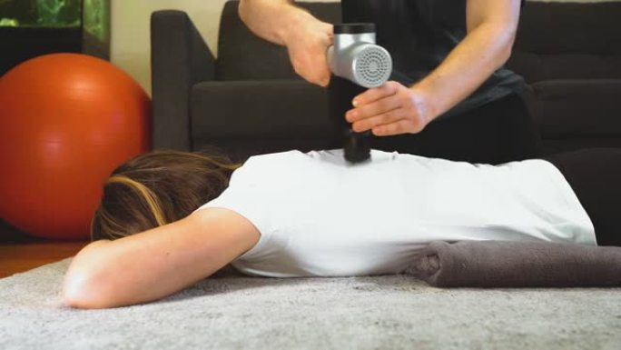 治疗师在家用按摩打击器按摩女人的背部。