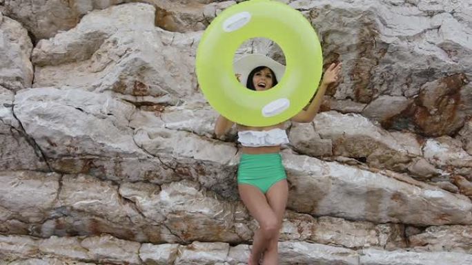穿着泳衣和帽子的迷人女孩在她的手上做了一个有趣的充气彩圈，并在岩石背景下看着相机。黄色充气浮子晒黑的