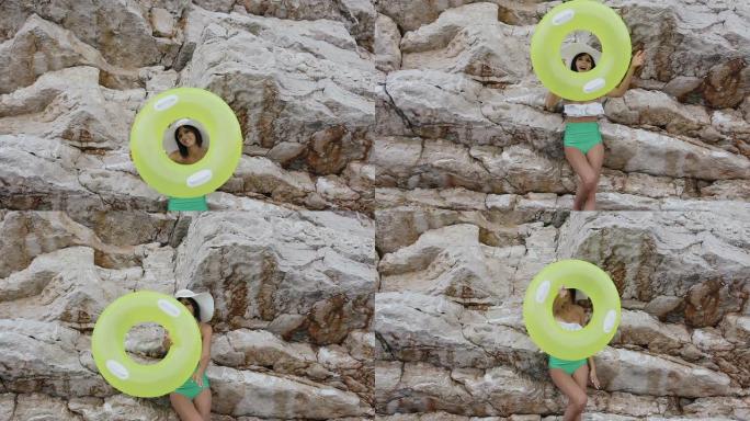 穿着泳衣和帽子的迷人女孩在她的手上做了一个有趣的充气彩圈，并在岩石背景下看着相机。黄色充气浮子晒黑的