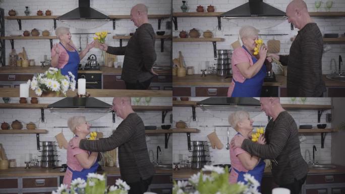 开朗成熟的高加索秃头男人给他可爱的妻子一束黄色郁金香。穿着粉红色t恤的金发白人妇女在厨房做饭时感到惊