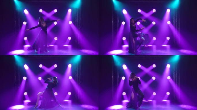优雅的年轻女子在黑暗的演播室跳舞。在烟雾中紫色的背光下，这位身材苗条的女演员流畅地举起双臂，跳着性感
