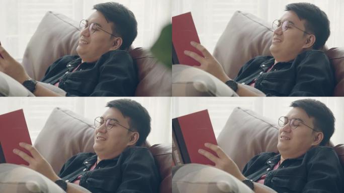 年轻的亚洲男子休息在沙发上看书和放松在家里的客厅