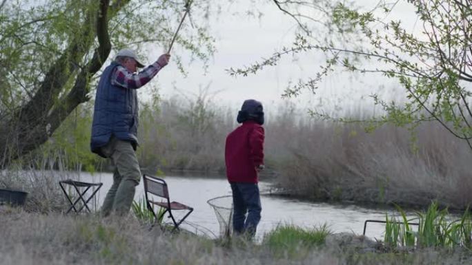 钓鱼时，爷爷和小男孩在乡间的芦苇和树木间休息时，用鱼竿在河里钓小鱼