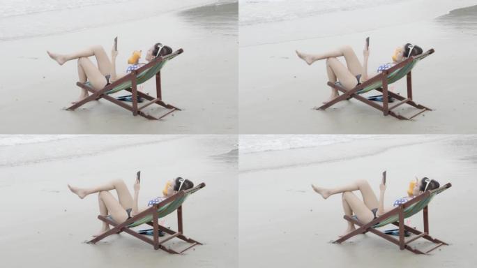 穿着比基尼性感的年轻亚洲女子在海滩热带度假时使用智能手机，女孩坐在躺椅上旅行，夏天在海边休闲时用手机