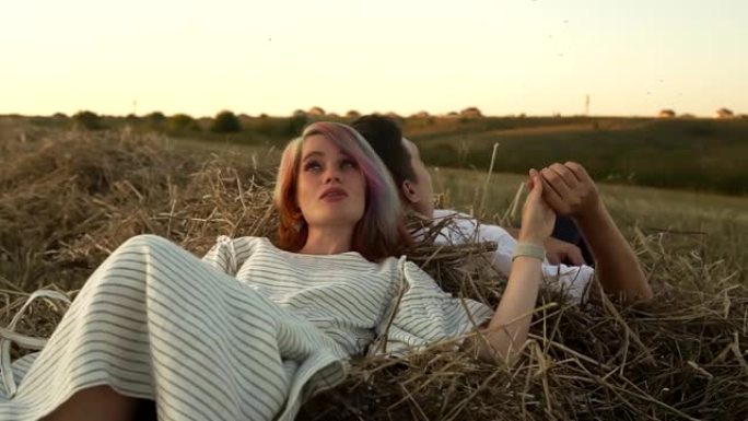 年轻的高加索夫妇躺在草堆上，牵着手在夏天的田野里聊天