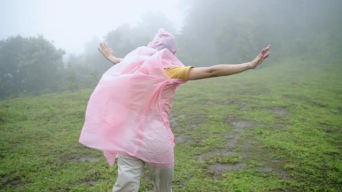 穿着粉色雨衣的年轻亚洲女性徒步旅行者的慢动作后视，在山中刮起大风，在雨中徒步旅行。漂亮女孩在美丽的大