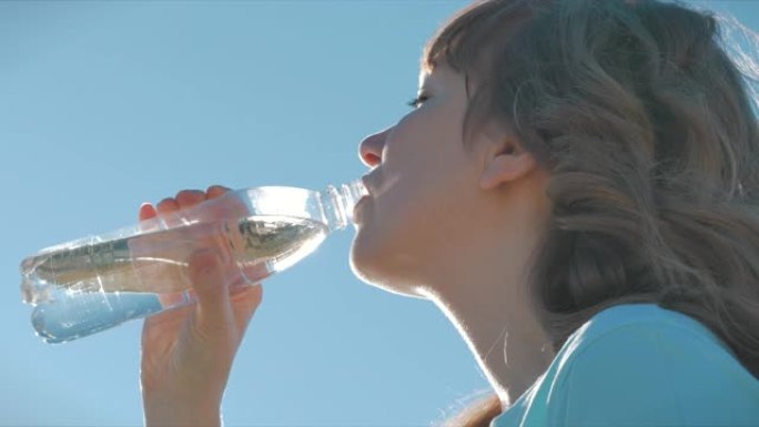 在露天的欧洲外表的女人在湛蓝的天空下从瓶子里喝水。健康的生活方式。