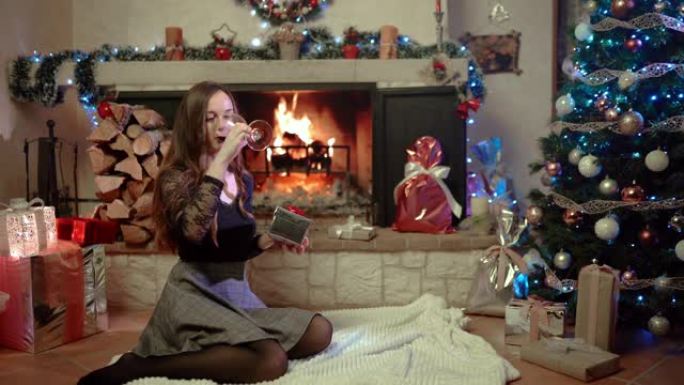 令人惊叹的圣诞节夜晚，在家里，有燃烧的壁炉和装饰的圣诞树，美丽优雅的女人坐在地板上，喝着红酒，手里拿