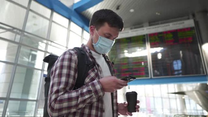 戴防护面罩和杯咖啡的男子使用智能手机在线办理登机手续，站在机场航站楼附近的出发到达时间表屏幕显示期间