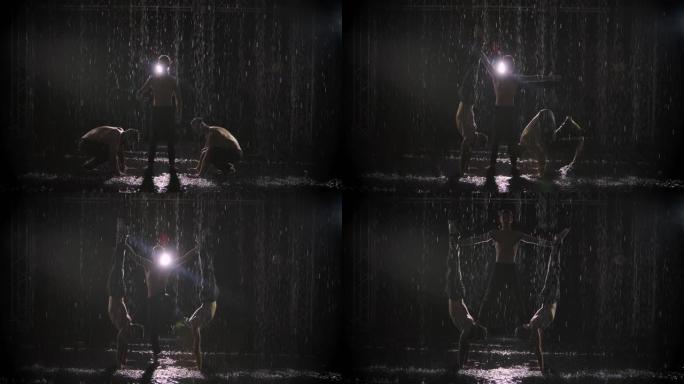 三个运动男子在雨中表演杂技特技的剪影。在黑色背景上聚光灯下的艺术家。慢动作