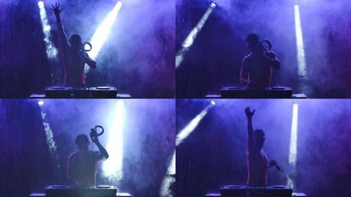 戴着耳机和墨镜的男性DJ的剪影在带有蓝光的黑暗烟熏工作室里用调音台控制器跳舞和混合音乐。晚上在雨中迪