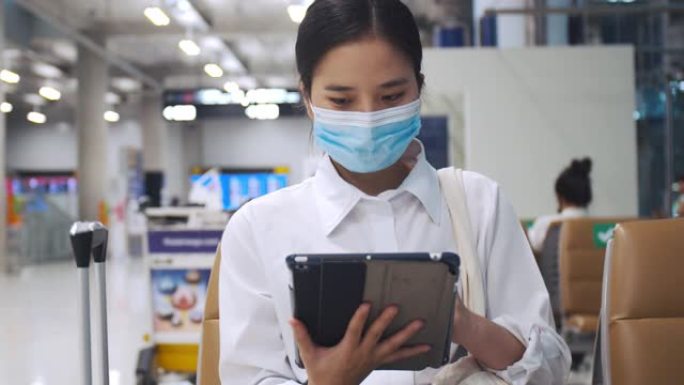 女商人戴着外科口罩保护装置，并在机场候机楼坐在椅子上使用平板电脑检查社交媒体供稿，并在新型冠状病毒肺