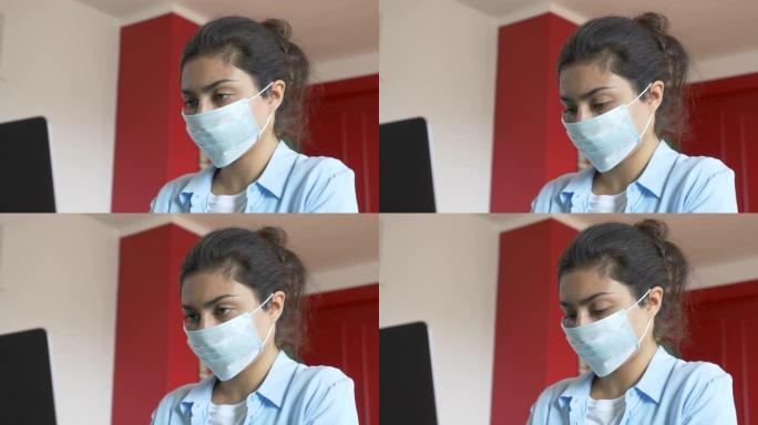一个戴着医用口罩的年轻印度妇女的特写镜头，使用笔记本电脑工作，公寓里明亮舒适的房间
