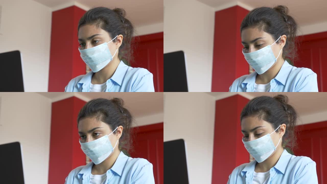一个戴着医用口罩的年轻印度妇女的特写镜头，使用笔记本电脑工作，公寓里明亮舒适的房间