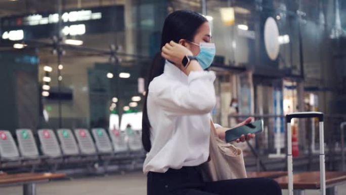 美丽的女性戴着外科口罩保护，并在机场航站楼外坐在椅子上使用智能手机，并在新型冠状病毒肺炎大流行期间保