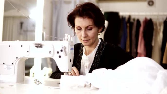 成年女性的肖像女裁缝在缝纫机上工作，穿着白色手工制作的衣服。裁缝工作室背景上的时尚服装。穿着婚纱的女