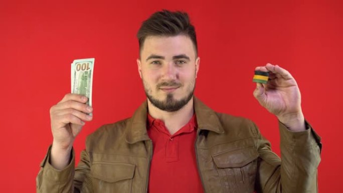 满意可爱的男人展示扑克筹码，并在红色背景上展示现金。手里拿着扑克筹码的扑克玩家。