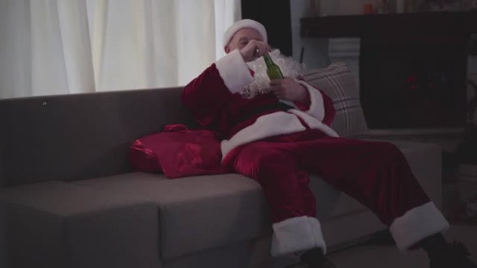 穿着圣诞老人服装的醉酒老人脸上留着假胡子坐在家里的沙发上，试图用牙齿打开瓶子。坏圣诞老人。酗酒、抑郁
