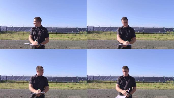 一名男子站在太阳能发电厂附近，手里拿着笔记本和笔。技术人员检查太阳能电池板的功能。替代能源服务质量控