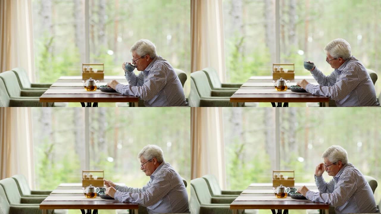 侧视中等的镜头是白发和戴眼镜的高级男子，他独自坐在咖啡馆的桌子上，喝茶和吃饭